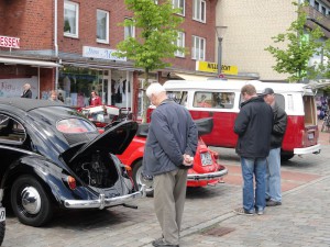 Drei Herren interessieren sich für zwei Oldtimer. Bei einem ist die Motorhaube hinten hochgeklappt und bietet einen Blick in das Innenleben.