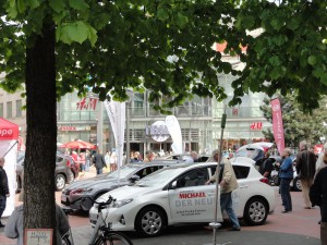 Ein Mann steigt in ein weißes Auto vor dem Tibarg Center. Im Hintergrund betrachten Passanten weitere Autos.