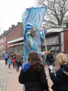 Eine Künstlerin mit fantasievollem Kostüm und Kopfbesatz in blau performt vor Zuschauern.