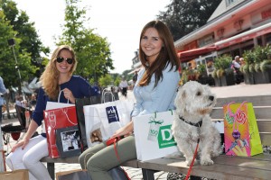 Das Bild zeigt zwei auf einer Bank sitzenden Frauen, die mit ihrem Hund zum Shoppen unterwegs sind.