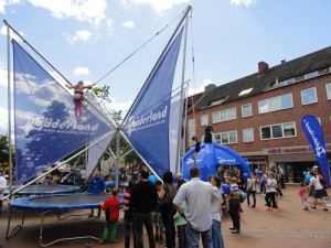 Kinder springen mit einem Gurt gesichert auf einem großen Trampolin und werden dabei von ihren Familien beobachtet.