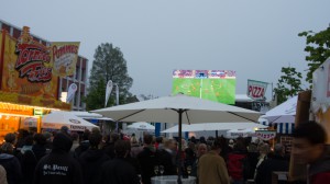 Über den Ständen hängt eine große Leinwand auf der Fußball gezeigt wird.