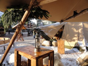 Das Foto zeigt den leicht verschneiten Weihnachtsmarkt-Platz im Tageslicht mit Blick auf das Tipi-Zelt. Auf einem Tisch im Vordergrund steht eine Öllampe.