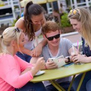 Ein Gruppe von vier Freunden sitzt an einem Café-Tisch und schaut gemeinsam auf ein Smartphone.